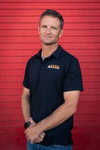 Man wearing a black polo shirt with an orange logo on the chest, standing with arms folded against a red wooden wall.