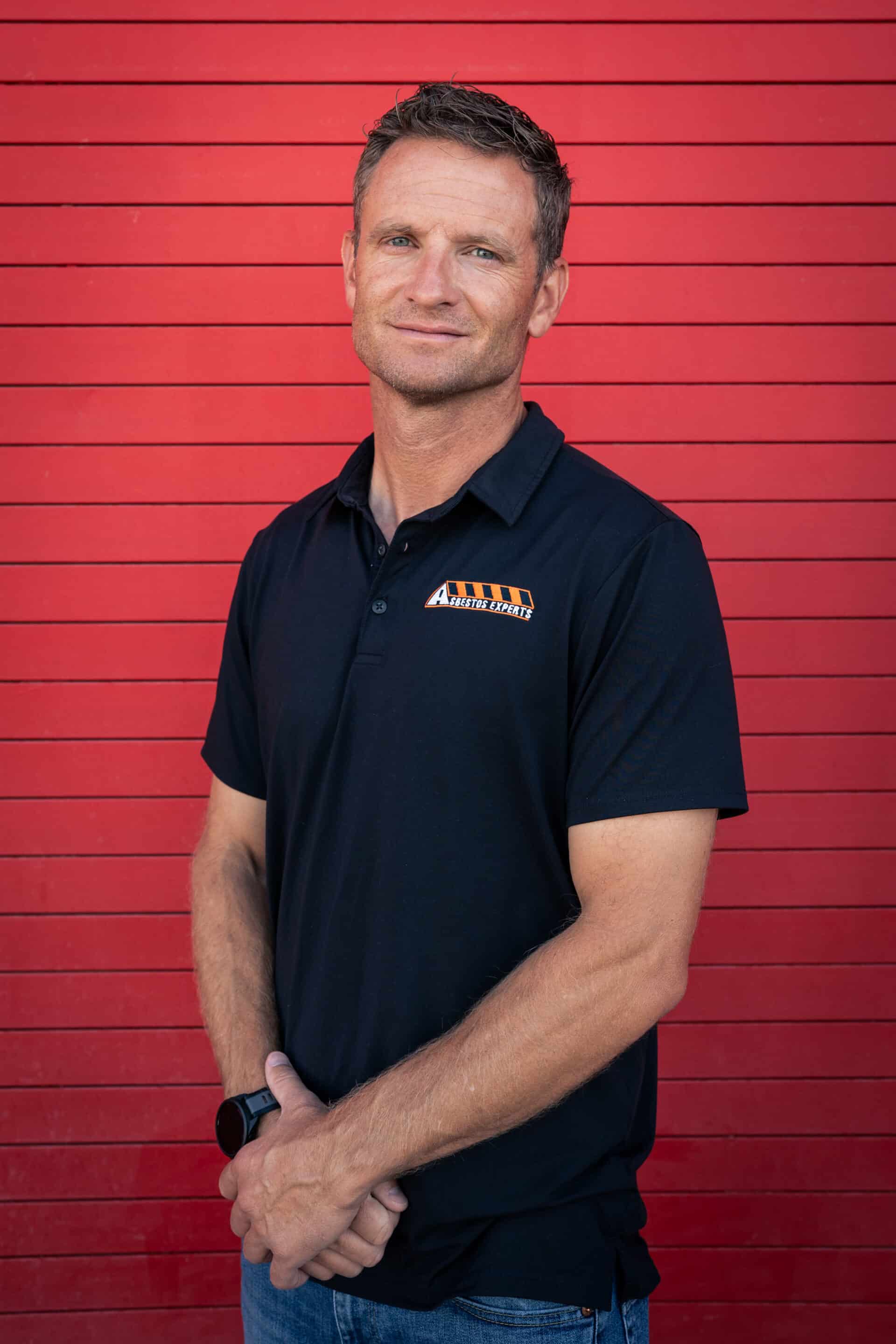 Man wearing a black polo shirt with an orange logo on the chest, standing with arms folded against a red wooden wall.