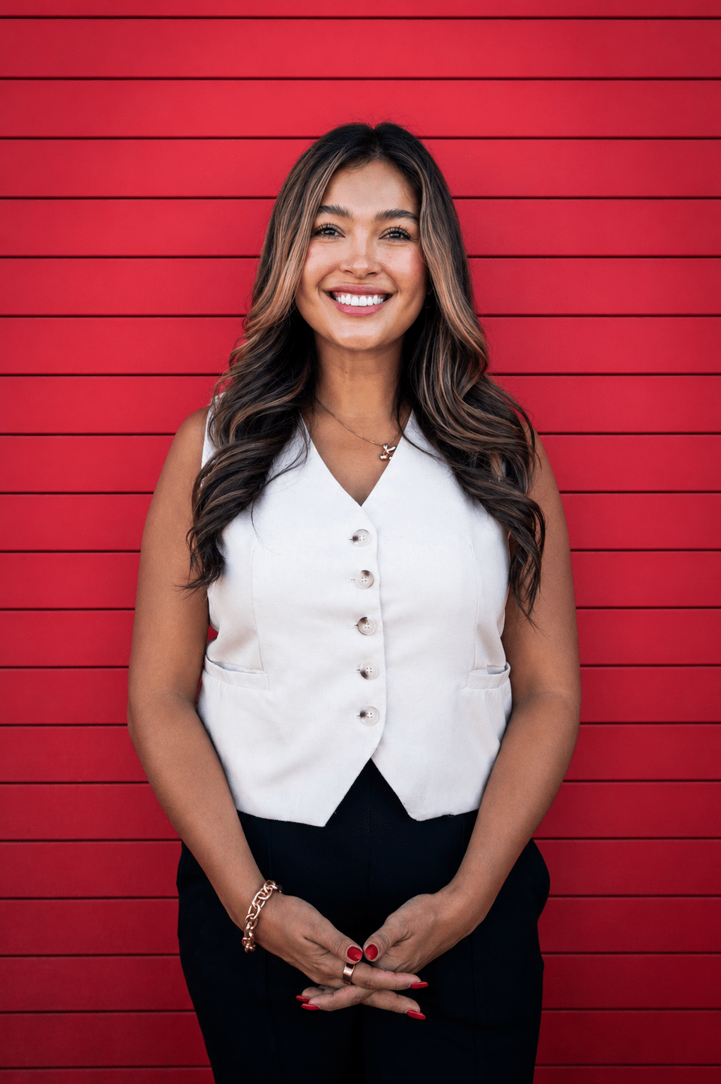Smiling woman with wavy hair in a white sleeveless vest and black pants, standing against a red slat wall.