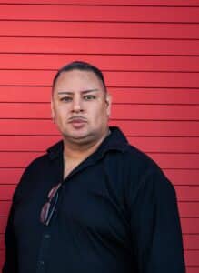 Portrait of a man with short dark hair and earrings wearing a black shirt against a red wooden wall in the background.