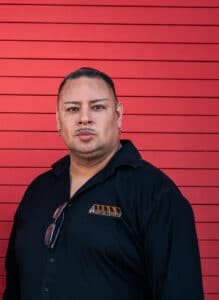 Portrait of a man in a black shirt with an asbestos experts logo, standing against a red horizontal-slatted wall.